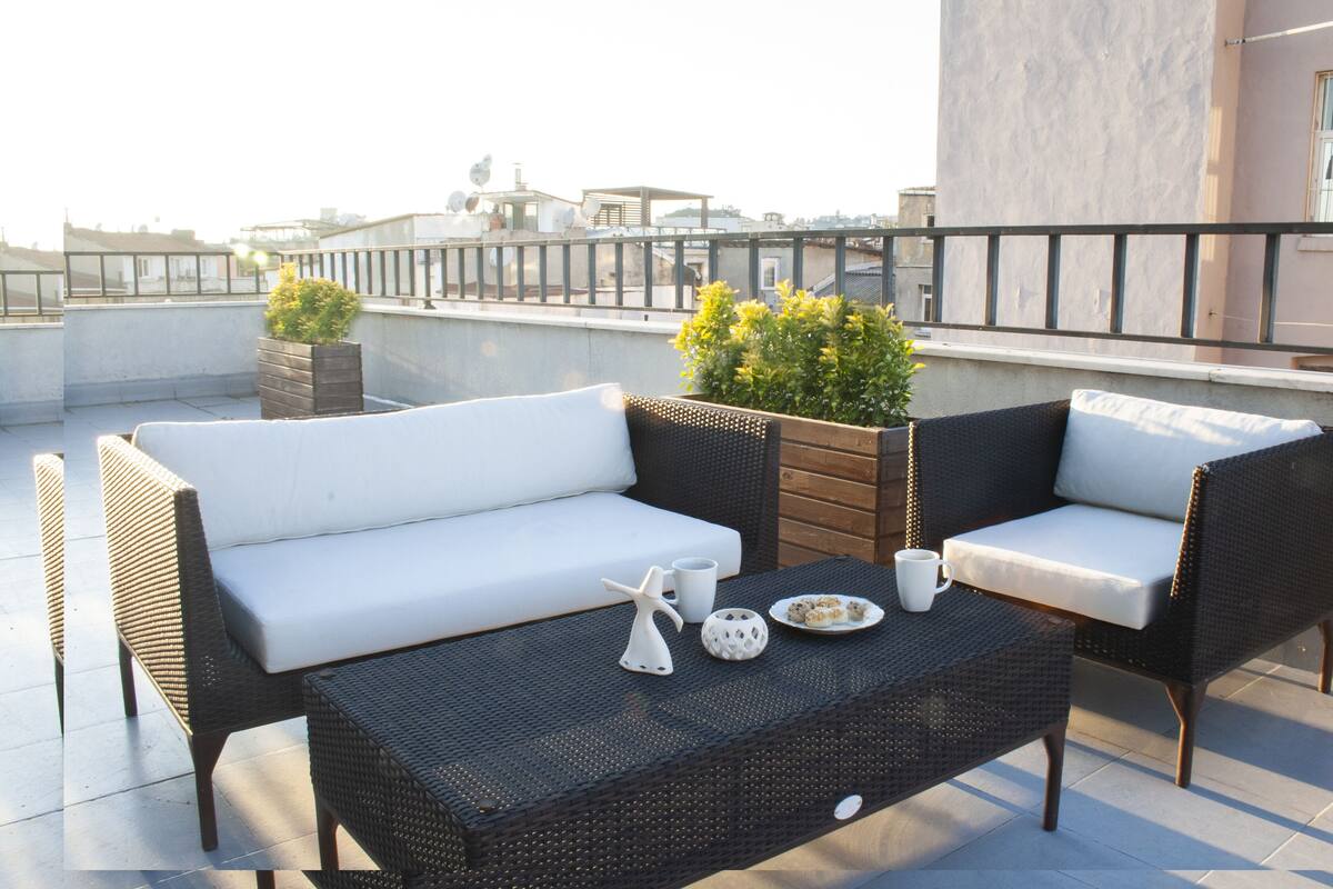 Una terraza en la azotea con mobiliario de mimbre, un sofá blanco con cojines y una pequeña mesa con objetos decorativos.