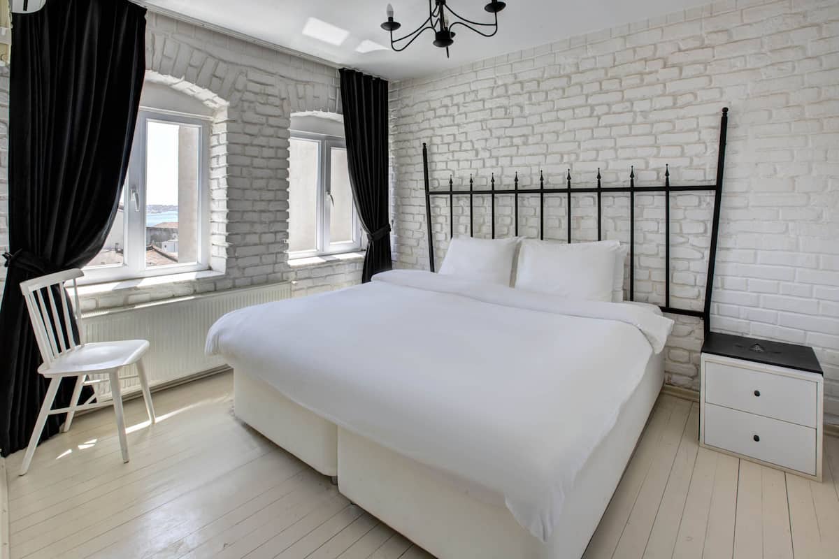 Un dormitorio con pared de ladrillo blanco, cama con ropa blanca, mesita de noche con cajones, una silla y una ventana con cortinas.