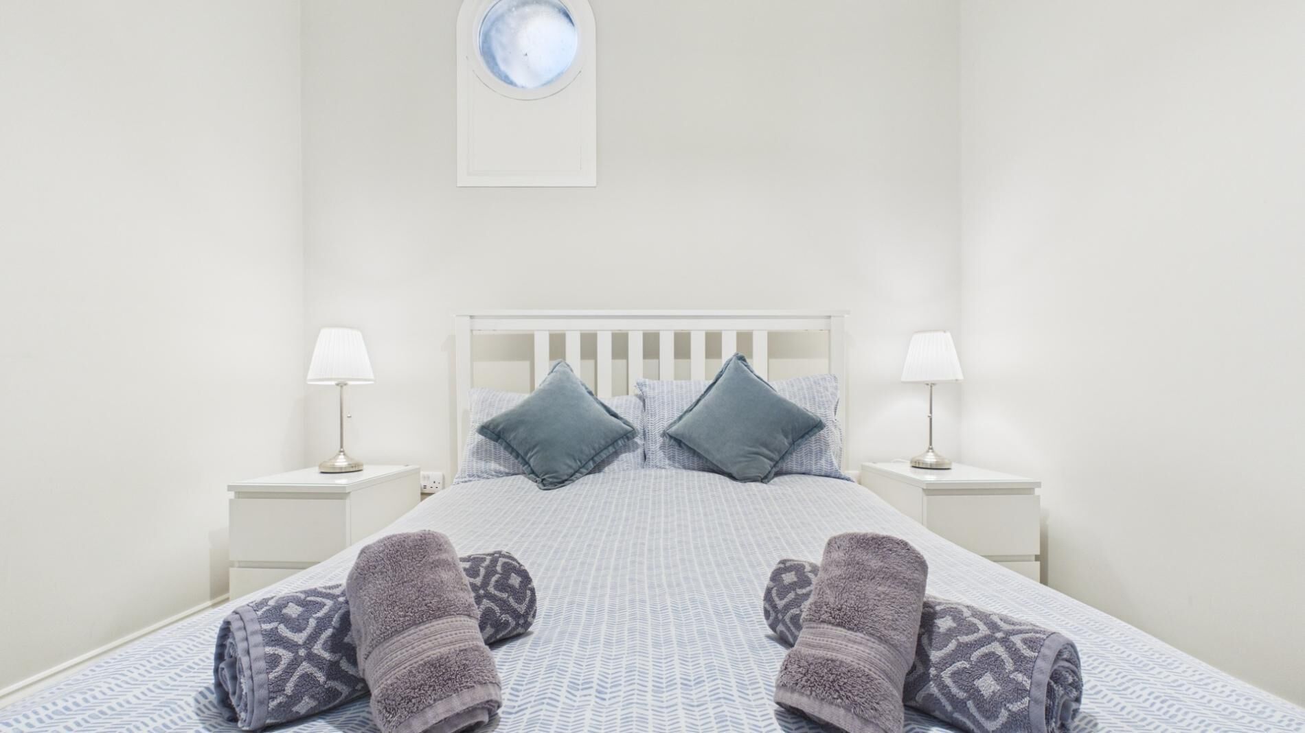 A white bedroom with a bed, two nightstands, two lamps, two pillows, and a round mirror on the wall.