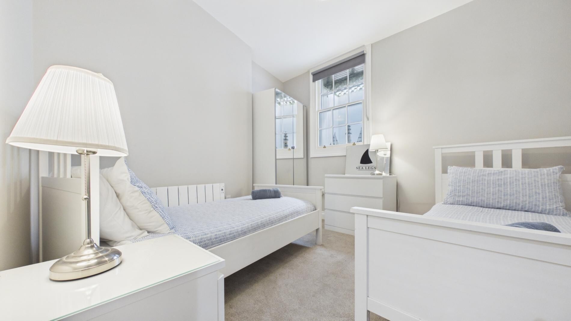 A bedroom with two beds, a bedside table with a lamp, a nightstand, a window, and a mirror.