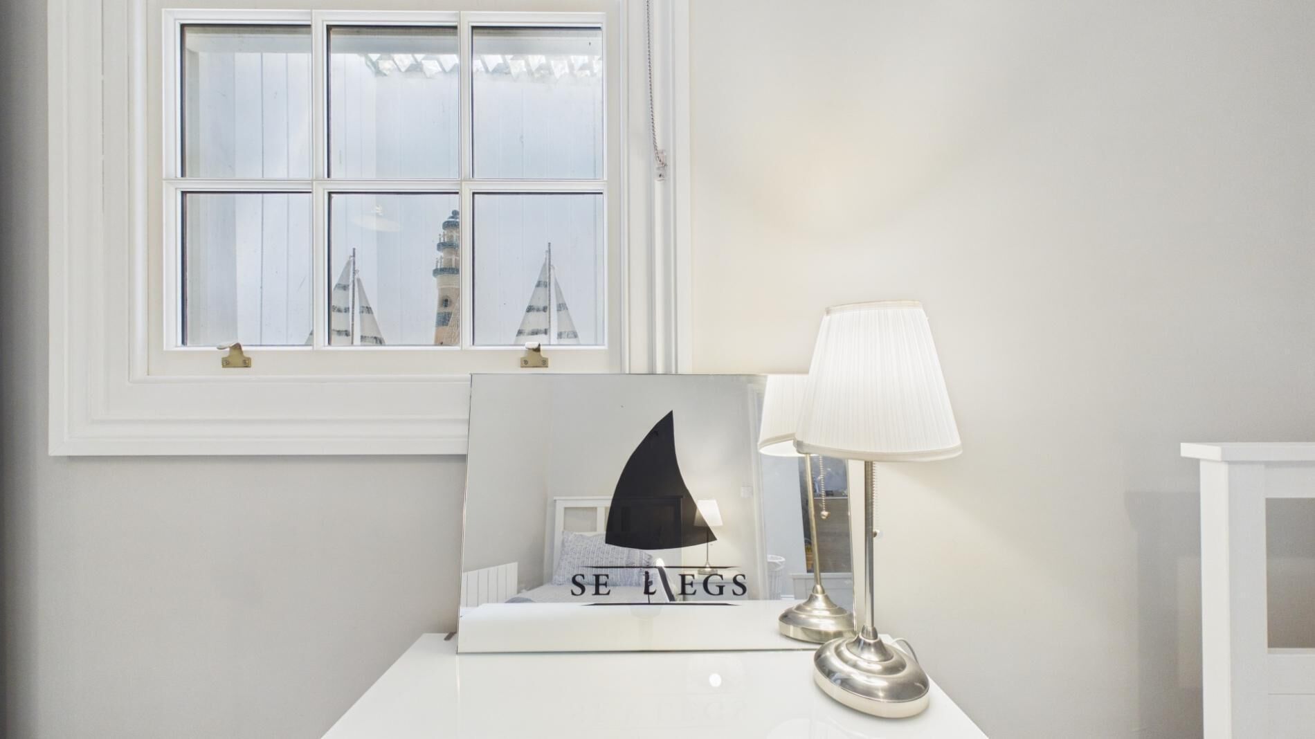 Een witte nachtkastje met een lijst met een afbeelding van een zeilboot en de tekst 'SE EGS', een lamp, en een raam met uitzicht op gebouwen.