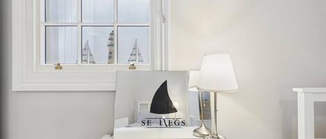 Een witte nachtkastje met een lijst met een afbeelding van een zeilboot en de tekst 'SE EGS', een lamp, en een raam met uitzicht op gebouwen.