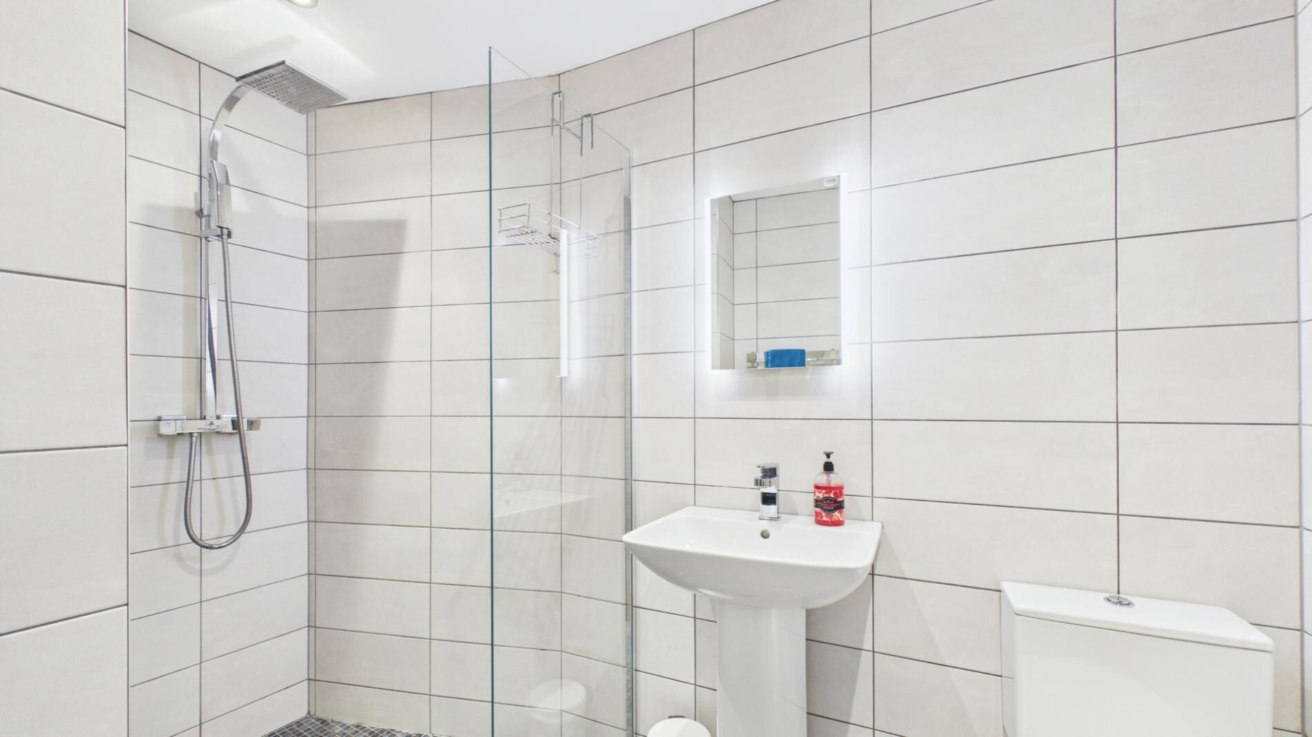 Un baño moderno con baldosas blancas, cabina de ducha de vidrio, lavamanos pedestal y un inodoro.