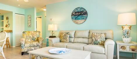 A living room with a light blue wall, a beige sofa, a patterned armchair, a coffee table with a decorative plate, a clock, and a lamp on a side table.