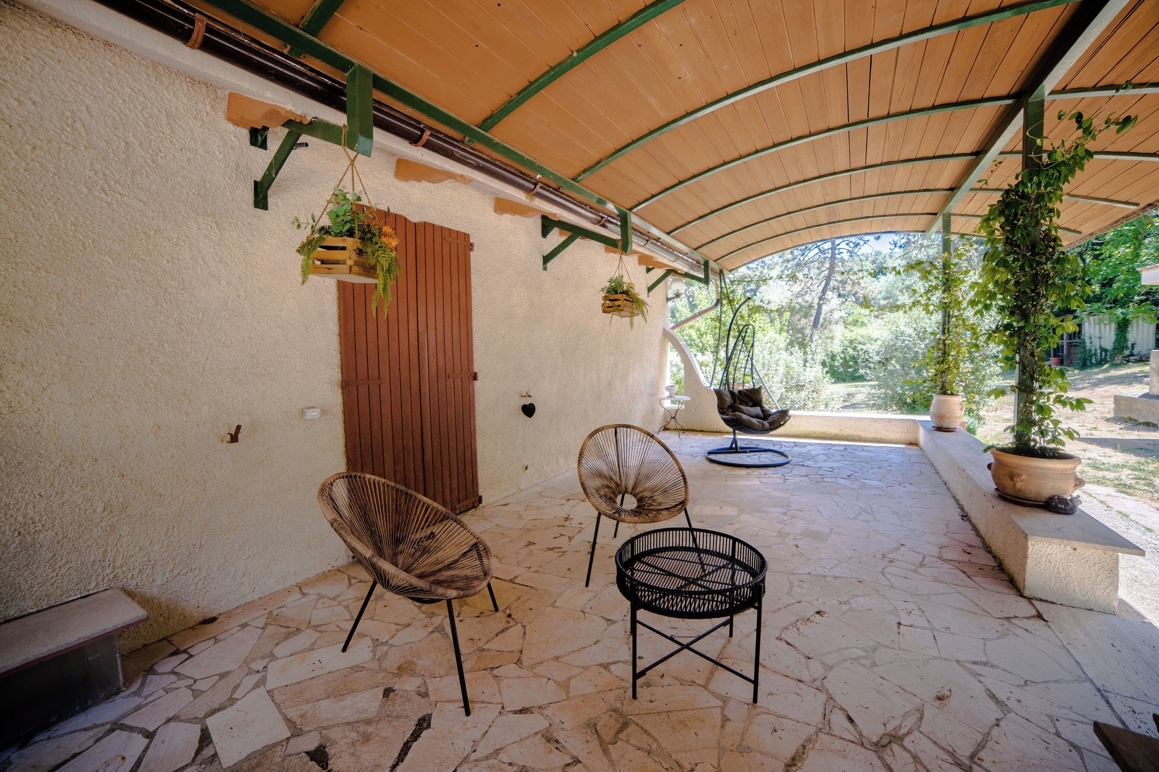 Een overdekte patio met stenen vloer, twee rieten stoelen, hangplanten en een houten deur.