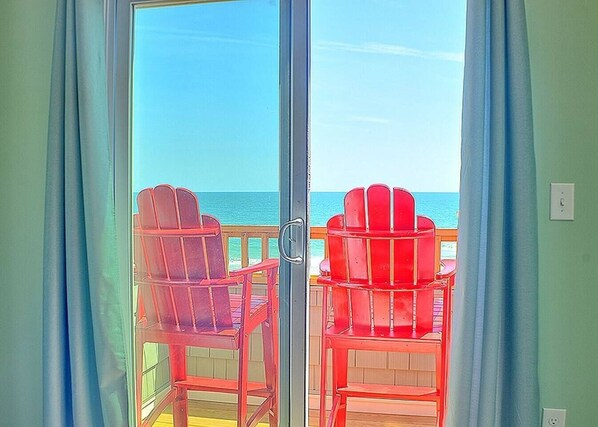 A view from a room with a sliding glass door leading to a deck with two red Adirondack chairs facing the ocean.