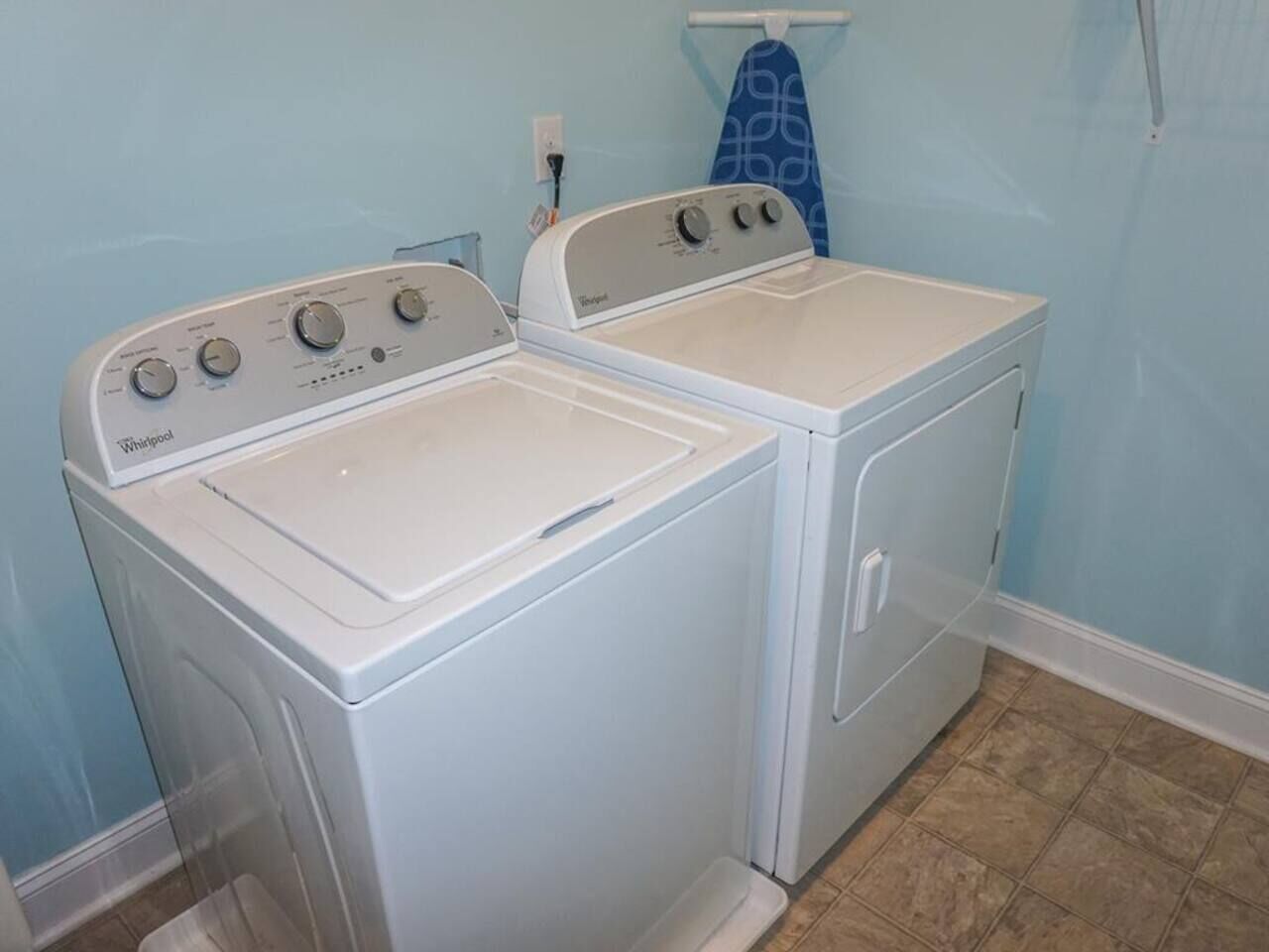 Une buanderie équipée de deux machines à laver et de deux sèche-linge Whirlpool, d’un porte-serviettes bleu et d’un sol carrelé.