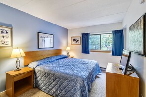 Une chambre à coucher avec un lit, deux tables de chevet avec des lampes, un bureau avec un ordinateur, une fenêtre avec des rideaux et des tableaux encadrés accrochés aux murs.