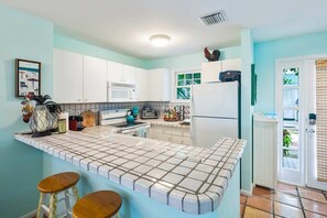 A kitchen with white cabinets, a tiled countertop, a microwave, a refrigerator, and a window with blinds.