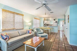 A living room with a sofa, a coffee table, a ceiling fan, and a kitchen in the background.