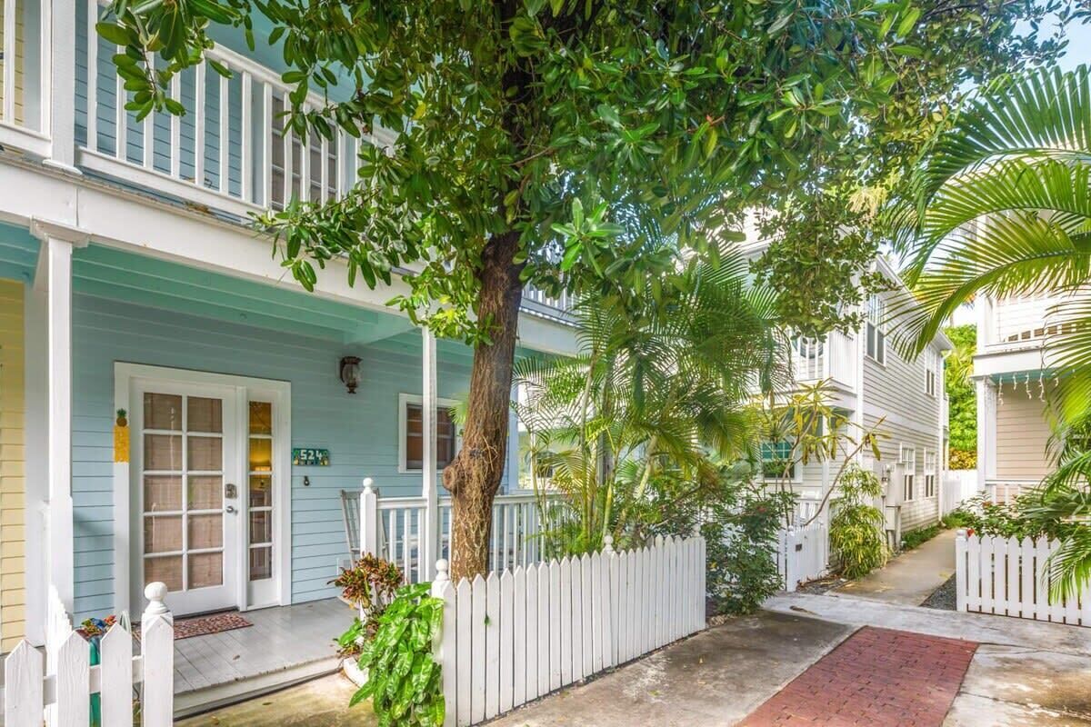 A two-story house with a white picket fence, a porch with plants, and a mailbox.