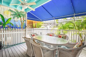 A covered outdoor patio with wicker furniture and a round table, surrounded by a white picket fence and lush greenery.