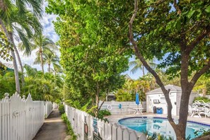 Uma piscina cercada por uma cerca branca de piquete, com árvores e um céu claro ao fundo.