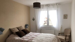 Ein Schlafzimmer mit einem Bett, einem Sessel, einem Fenster mit Vorhängen und einer Wandlampe.
