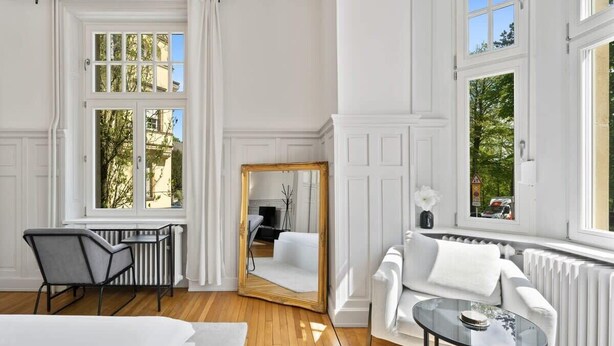 Ein heller Raum mit weißen Wänden, einem großen Spiegel, einem weißen Sofa, einem kleinen Tisch und einem Fenster mit Blick auf Bäume.