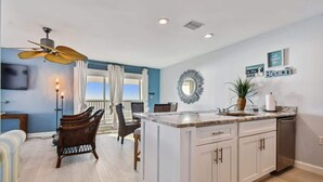 A kitchen with a central island, white cabinets, and a ceiling fan. There is a dining area with chairs and a table, and a view of the outdoors through a window. - 3Bedroom Beachfront Condo-Gulf View-On the Sand! (Indian Rocks Beach)