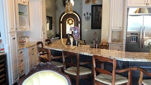 A kitchen with white cabinetry, a marble island, wooden chairs, and a large chandelier.