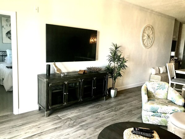 Ett vardagsrum med en platt-TV som är monterad på väggen, en mörk träinredd TV-möbel, en krukväxt och en blommönstrad soffa.