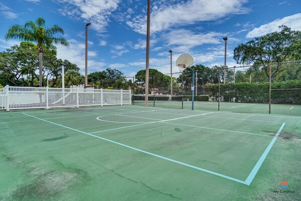 Una cancha de tenis al aire libre con valla blanca, aro de baloncesto y árboles de fondo.