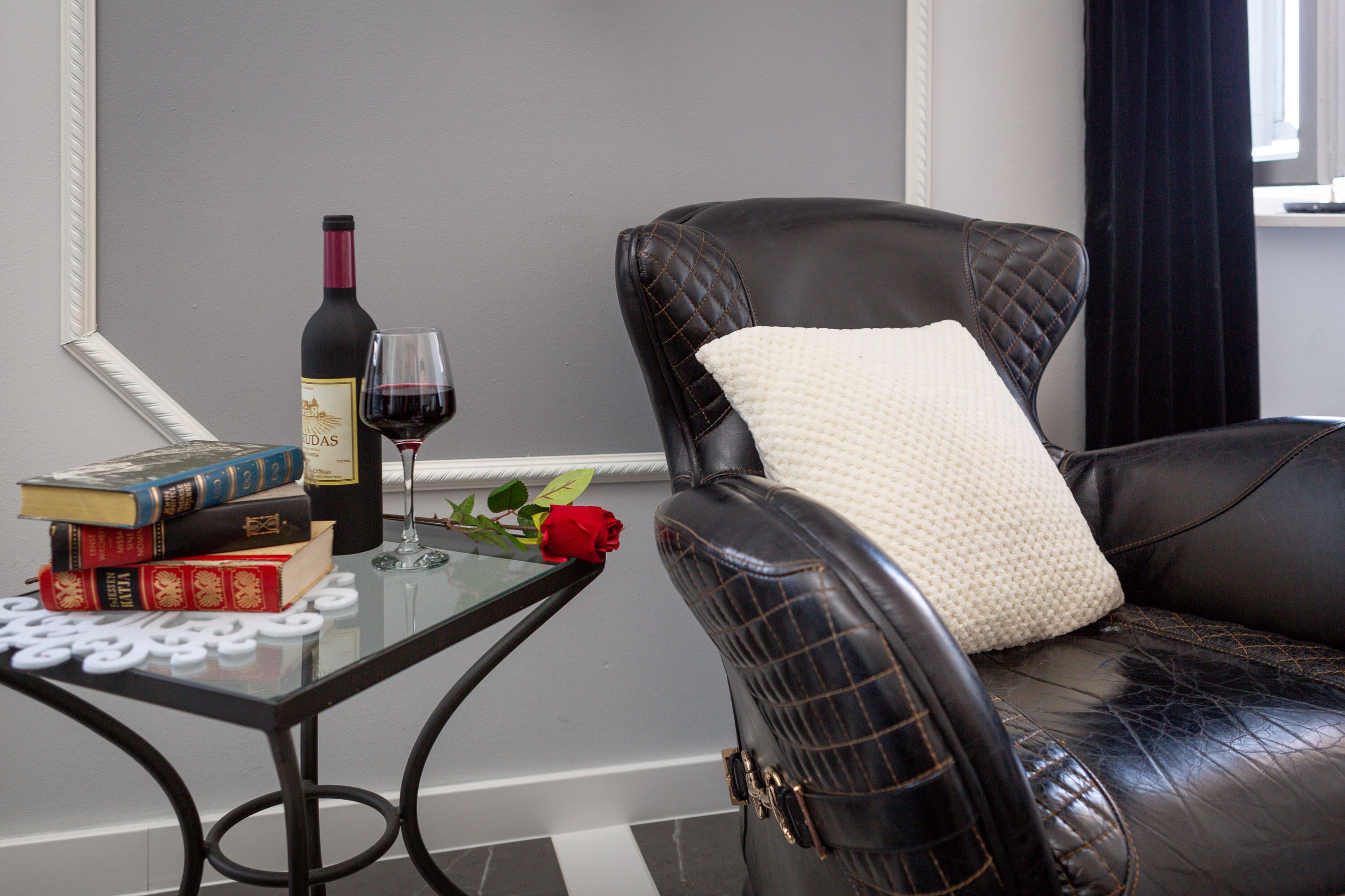 Un sillón de cuero con un cojín blanco, una mesa de vidrio con una botella de vino y libros, y una rosa roja.