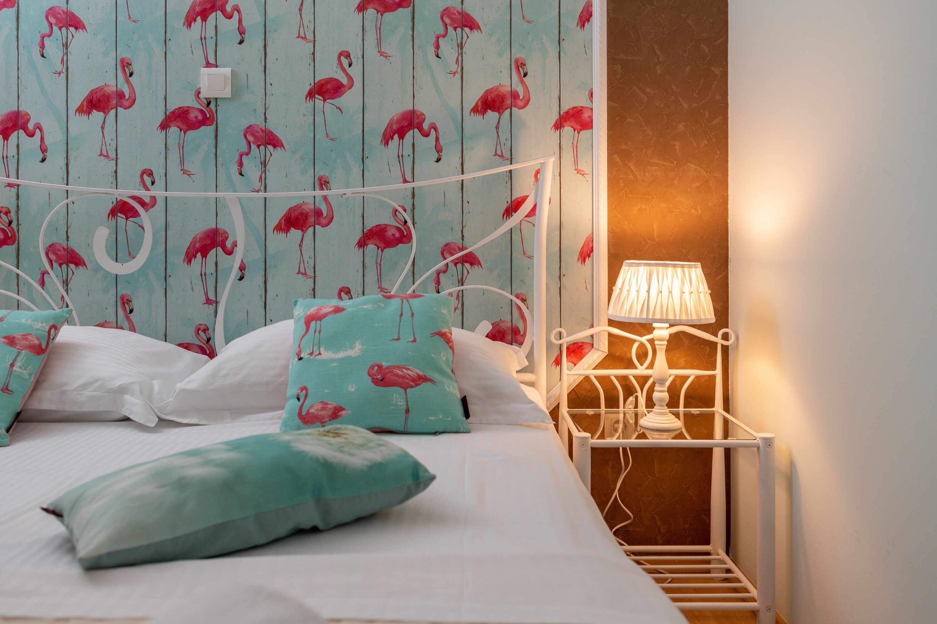 Un lit avec des oreillers blancs et turquoise, une tête de lit en métal blanche et un mur orné d’un motif de flamants roses.