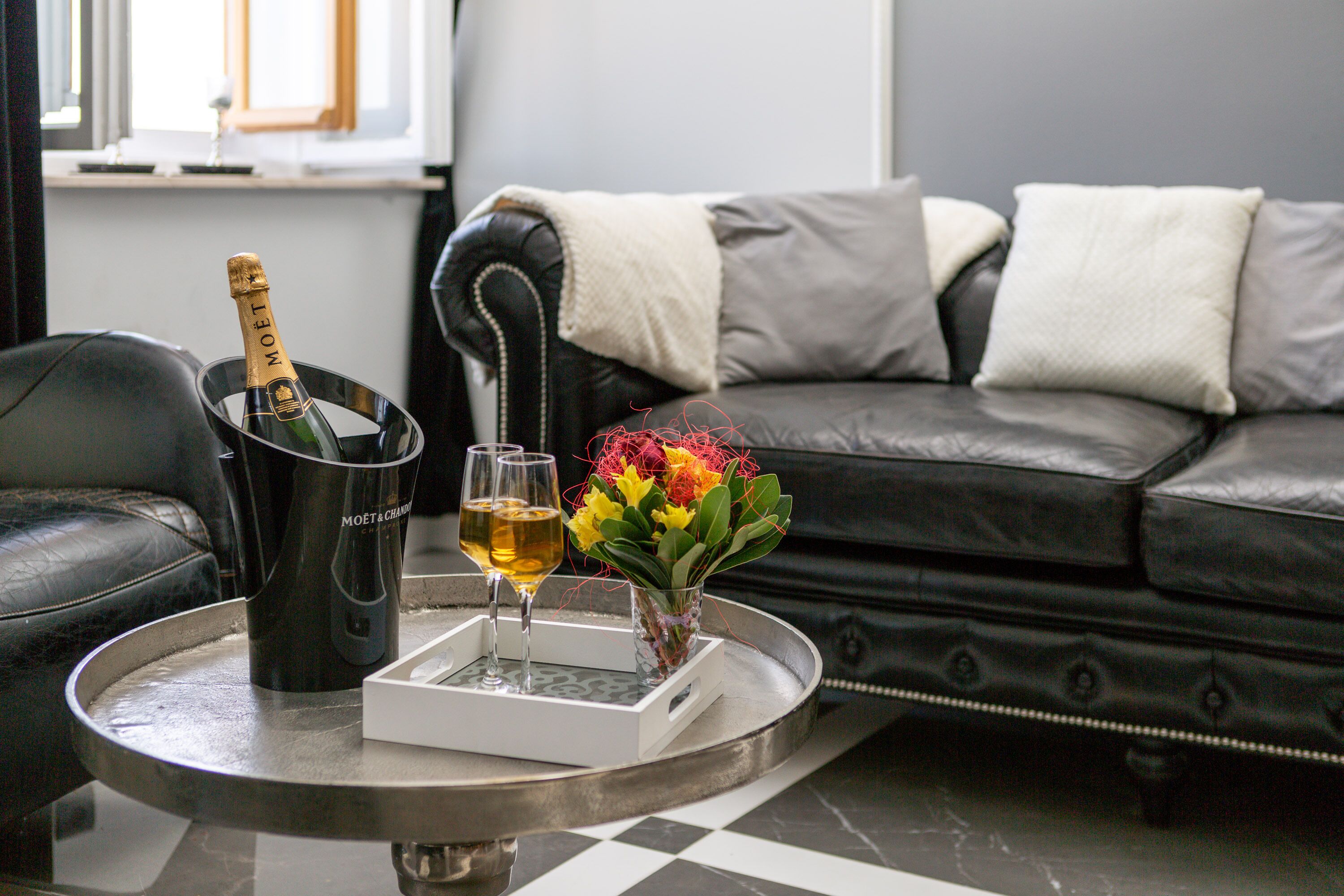 A black leather sofa with a white throw blanket, a tray with a bottle of champagne, two glasses, and a vase with flowers.