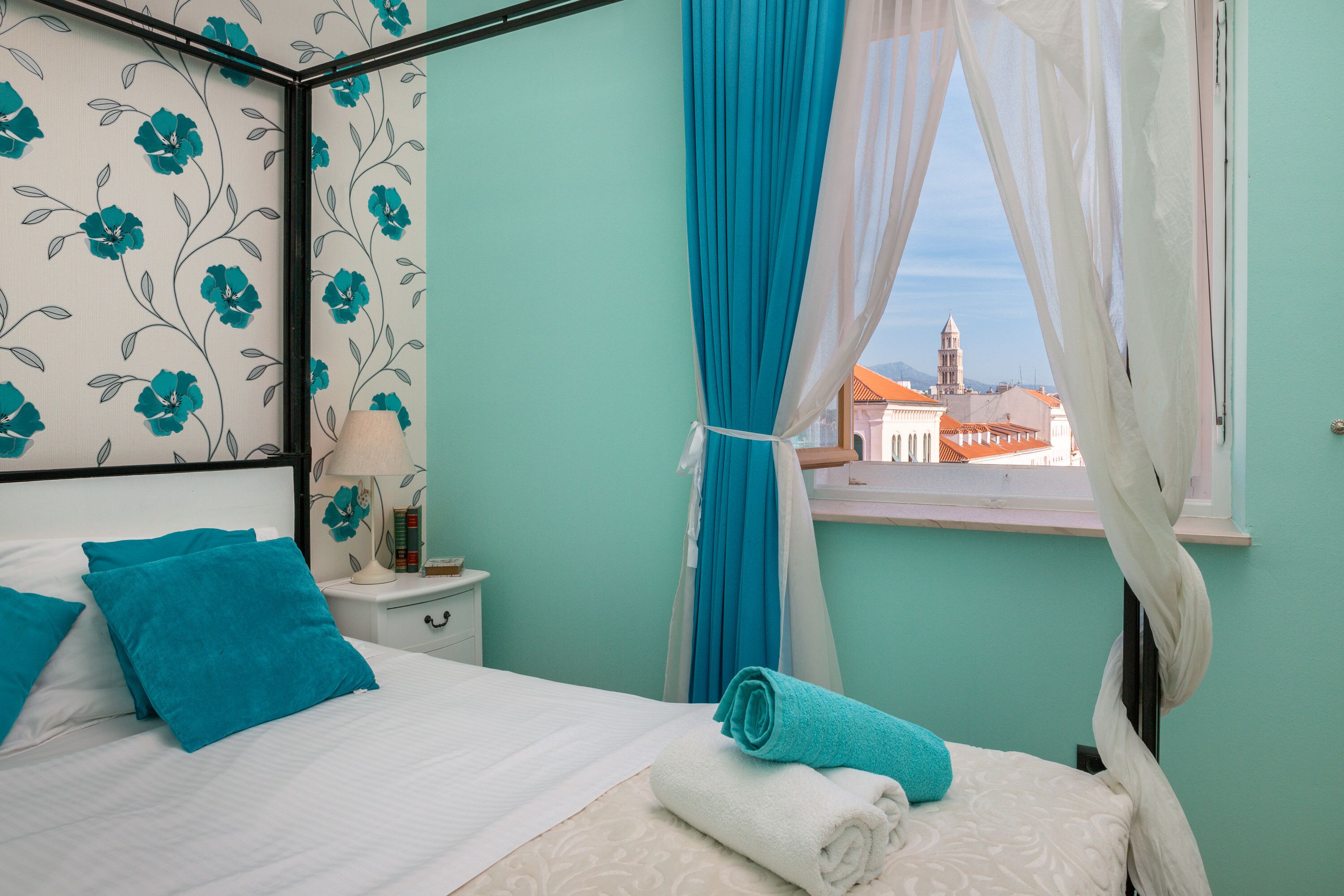 A bedroom with a bed, a nightstand, a window with curtains, and a view of buildings outside.
