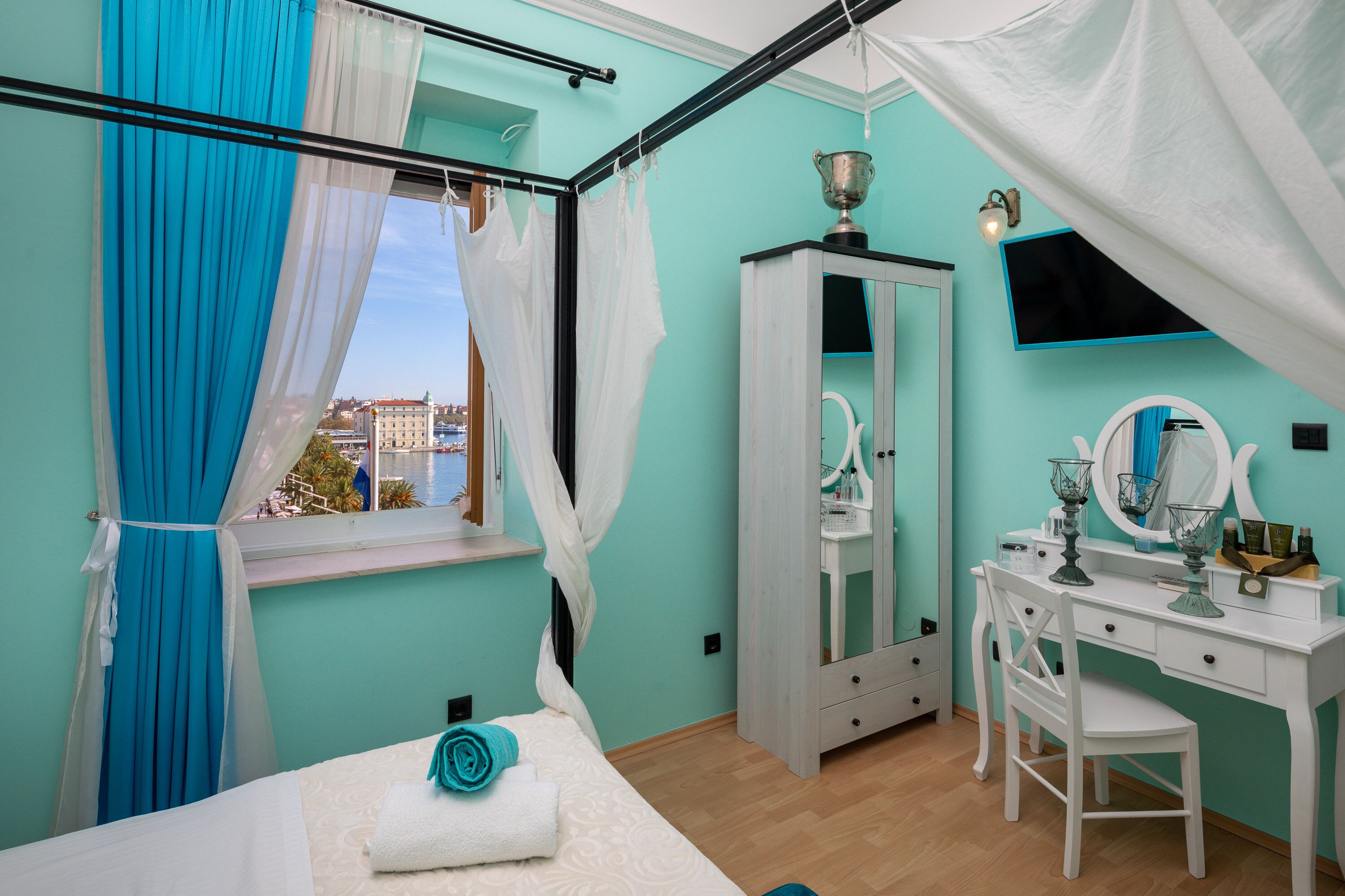 A bedroom with a bed, a vanity with a mirror, and a view of buildings through a window.