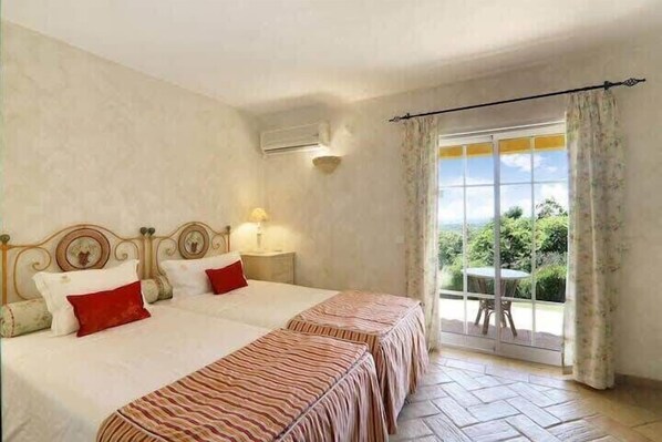 A double bed with a striped bedspread, a bedside table with a lamp, and a view of greenery through a window with curtains. - Villa Faustine, huge lawn, sea view, kid play area (Boliqueime)