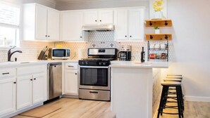 A modern kitchen with white cabinets, a stainless steel dishwasher, a microwave, an oven, and a coffee maker. There is a wooden shelf with a vase and a window with a view of the outside. - Staycation House | Modern and Spacious (Austin)