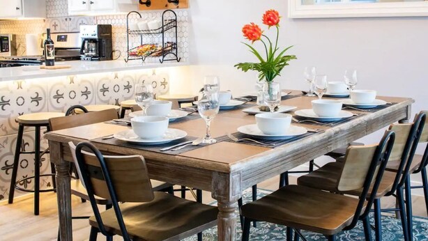 A dining area with a wooden table set for six, surrounded by chairs. A kitchen is visible in the background with modern appliances and a coffee machine. - Staycation House | Modern and Spacious (Austin)