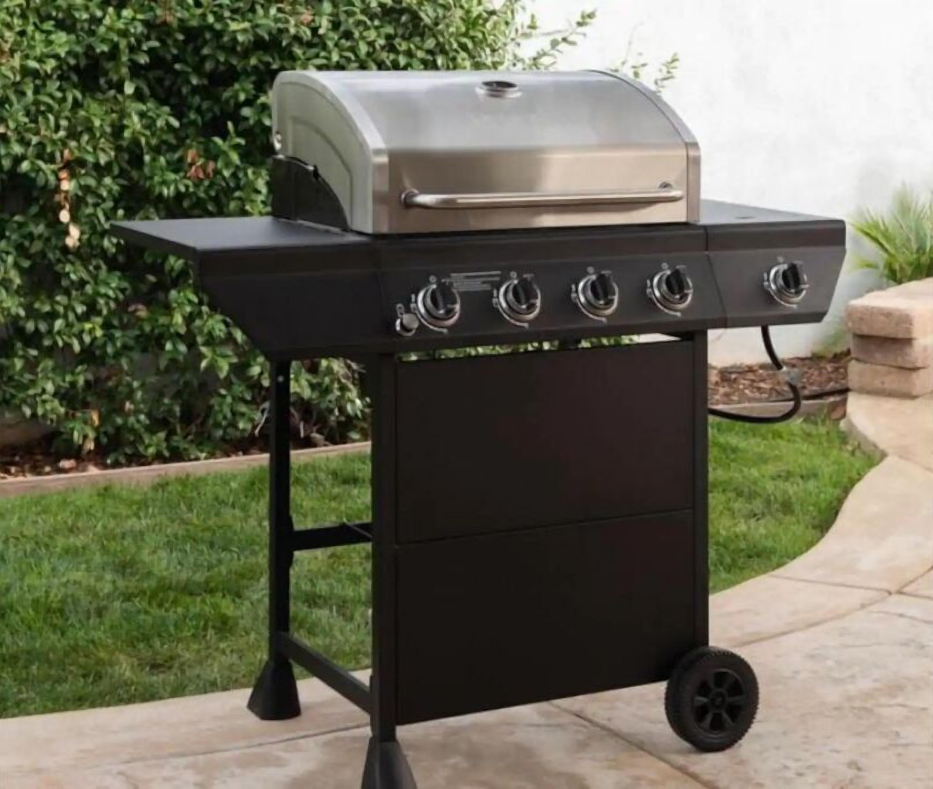 A portable barbecue grill with four burners and a stainless steel lid, placed on a patio with a grassy area and a hedge in the background.