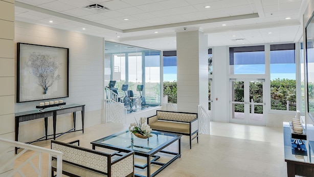 A spacious, modern lobby with a glass wall offering a view of the outdoors, a seating area with a glass coffee table, and a framed artwork on the wall. - Beachfront Condo With Pools, Tennis Courts and gym (Marco Island)