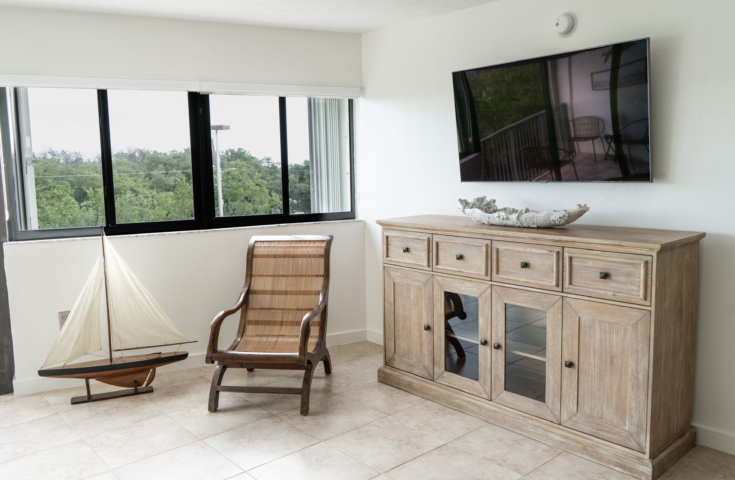 A room with a large window, a sailboat model, a wooden chair, a cabinet with drawers, and a flat-screen TV mounted on the wall.