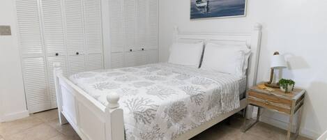 Un dormitorio con cama, armario blanco y mesita de noche con lámpara y planta.