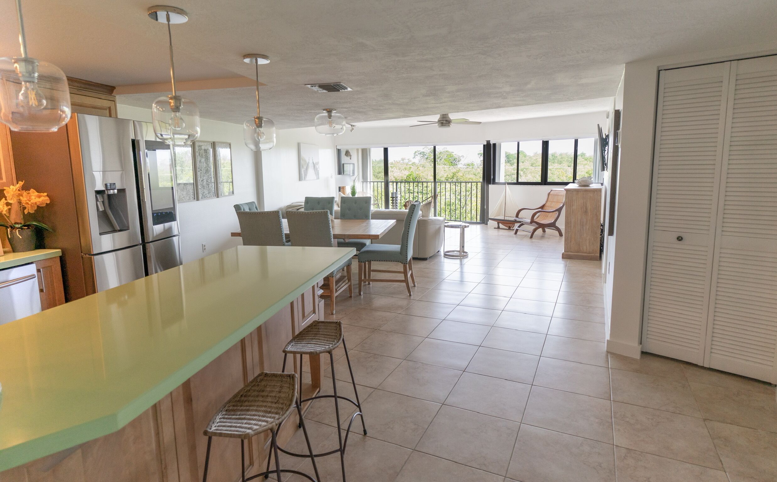 A spacious kitchen with a long island, bar stools, and a dining area with chairs. There is a refrigerator, a water dispenser, and a ceiling fan. The room has large windows and a view of the outdoors.