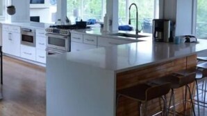 A modern kitchen with a large island, wooden ceiling, and large windows.