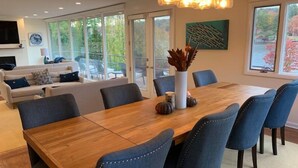 A spacious dining area with a wooden table and blue upholstered chairs, a white vase with decorative items, and a large window with a view of trees and a building.