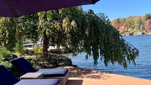 A deck with lounge chairs and a large umbrella, overlooking a lake with trees and a dock in the background.