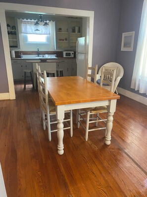 A dining area with a wooden table and chairs, a kitchen in the background, and a chair with a sign. - Cozy Bungalow located 2 blocks from the beach-  (Galveston)