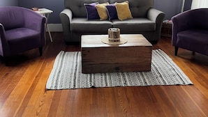 A living room with a grey sofa, two purple armchairs, a wooden coffee table, and a patterned rug on wooden flooring. - Cozy Bungalow located 2 blocks from the beach-  (Galveston)