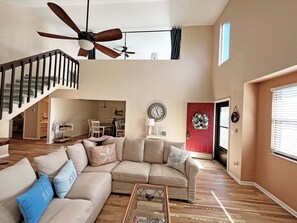 A spacious living room with a high ceiling fan, a staircase, a large beige sectional sofa, and a dining area in the background. - 3 Sabor de Sal-Seas The Day (St. Augustine)