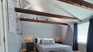 A bedroom with a bed, bedside tables, a bench, a patterned rug, wooden flooring, and exposed wooden beams. - Modern Condo in the West End, central to everything you want to enjoy in P-Town. (Provincetown)