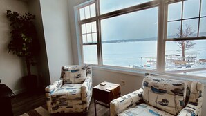 Una sala de estar con dos sillones estampados, una mesita, un ventanal con vistas a una masa de agua y una planta en maceta.
