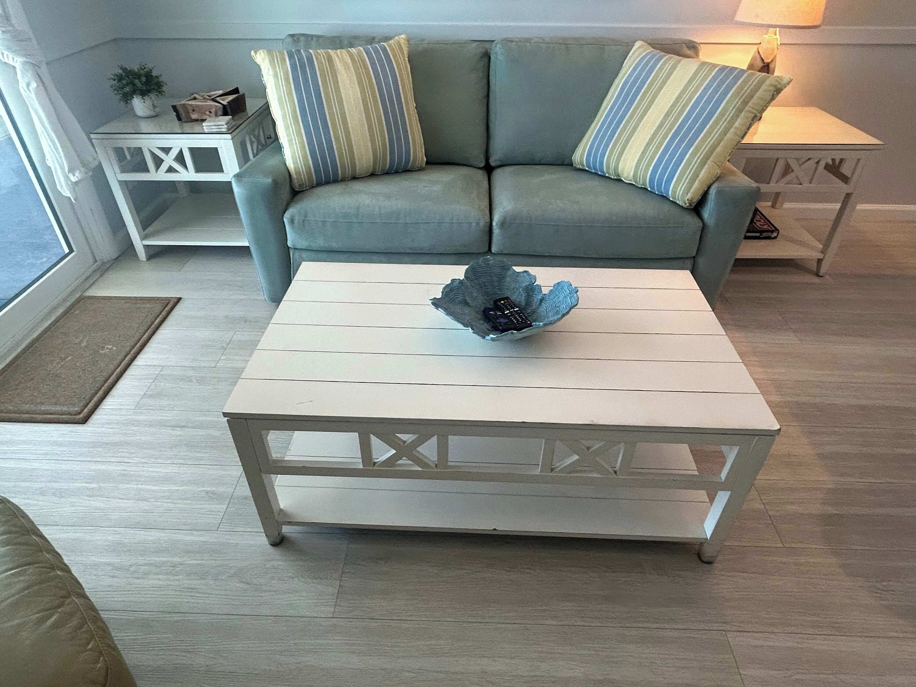 A living room with a grey sofa, striped pillows, a white coffee table with a decorative bowl, and a small white side table with a lamp.