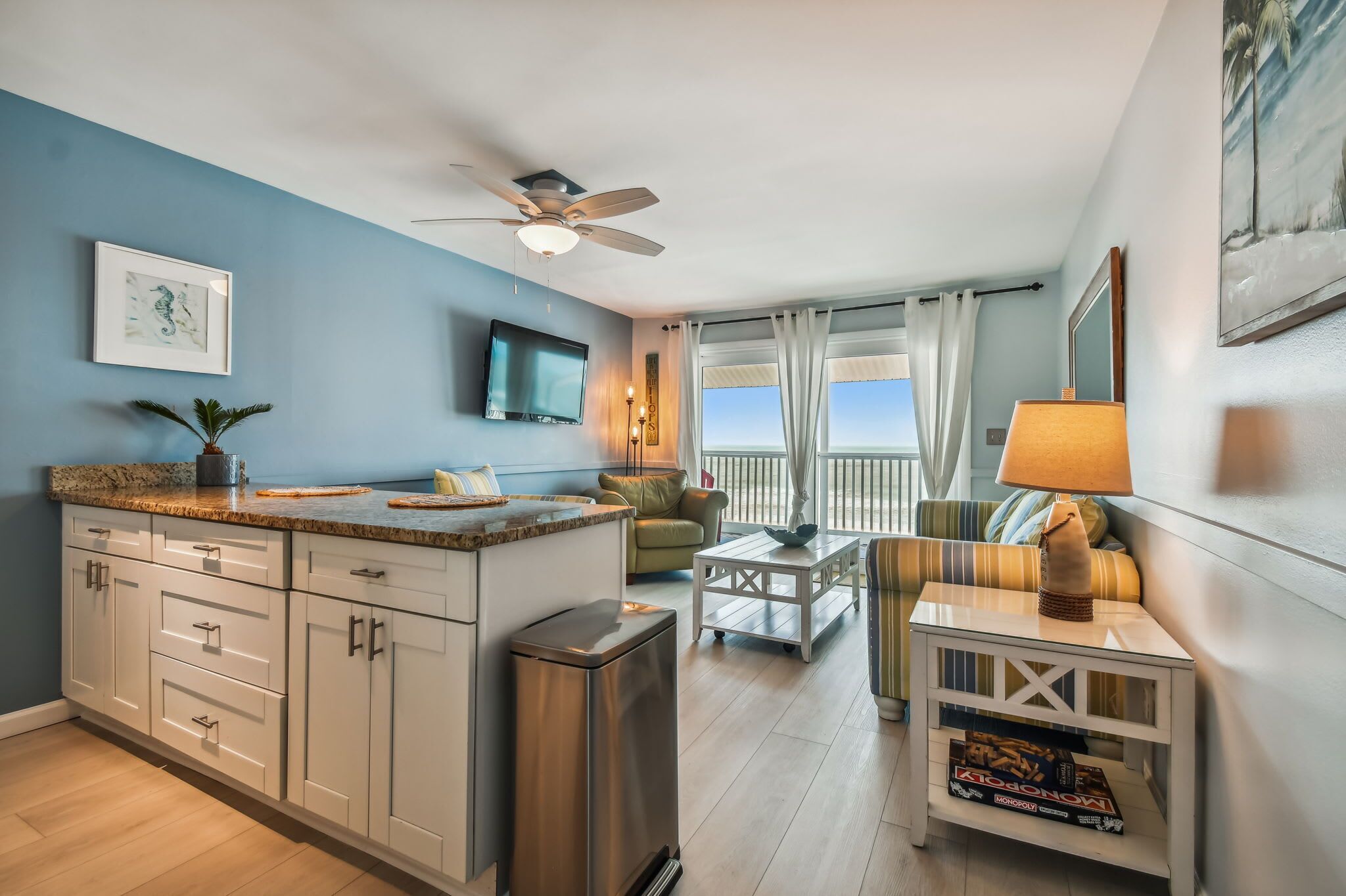 A modern kitchen with a granite countertop, white cabinets, and a stainless steel trash can. There is a dining area with a striped sofa and a table, and a balcony with a view of the ocean.