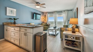 A modern kitchen with a granite countertop, white cabinets, and a stainless steel trash can. There is a dining area with a striped sofa and a table, and a balcony with a view of the ocean.