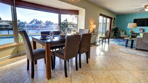 A dining area with a view of a body of water, a dining table with chairs, a lamp, and a TV in the living area. - Wildlife Views at CocoaBeachRetreat WaterfrontOasis,PrivatePool&WildlifeOnCanal! (Cocoa Beach)