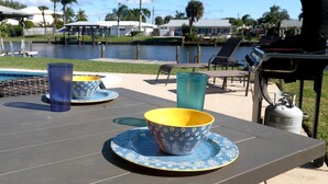 A dining table with two patterned bowls and a blue glass, set outdoors with a pool and palm trees in the background. - Wildlife Views at CocoaBeachRetreat WaterfrontOasis,PrivatePool&WildlifeOnCanal! (Cocoa Beach)