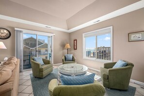 A spacious living room with a round glass coffee table, two green armchairs, a beige sofa, and a large window offering a city view. - Whispering Sands - 3Br ,max occ 10, midtown, ocean view, pool (Ocean City)